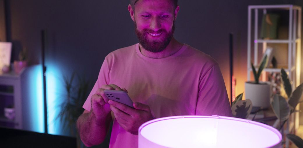 Mężczyzna steruje inteligentnym oświetleniem za pomocą smartfona, zmieniając kolor światła lampy w nowoczesnym wnętrzu smart home.